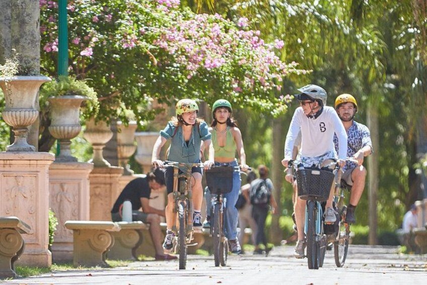 People riding bicycles on a paved path in a park with stone planters and flowering trees. 2