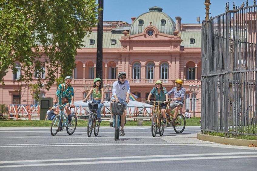 A group of cyclists on a street with a large pink building in the background. 1