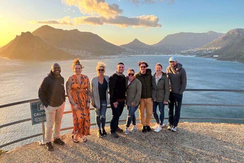 En grupp människor står på en stenig strand med en vattenyta och berg i bakgrunden. 2
