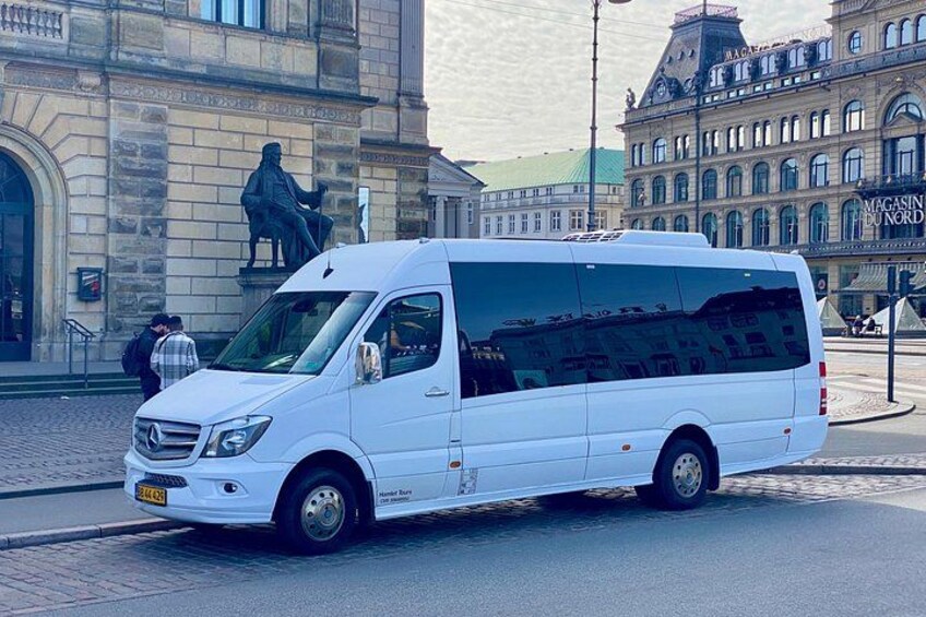 En hvid varevogn parkeret på en brostensbelagt gade foran en historisk bygning med en statue. 2