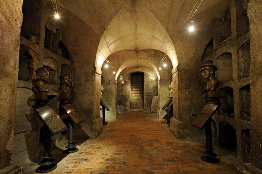 An underground passageway with arched ceilings and stone walls, featuring busts and plaques along the sides. 1