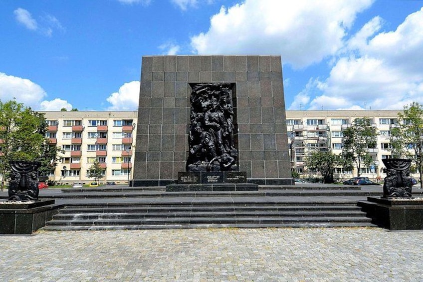 A large black monument with a relief sculpture, surrounded by steps and trees, with apartment buildings in the background. 2
