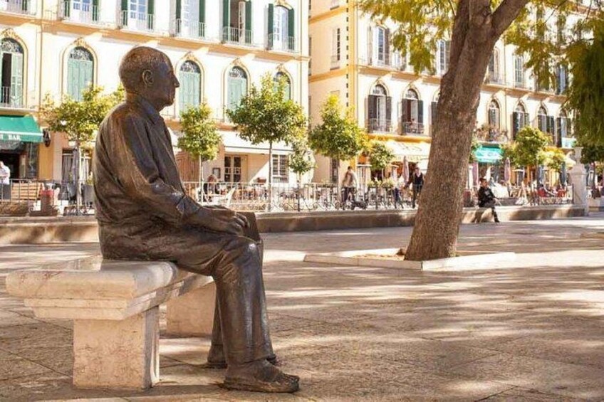 Una estatua de bronce de un hombre sentado en un banco en una plaza pública, con edificios al fondo. 2