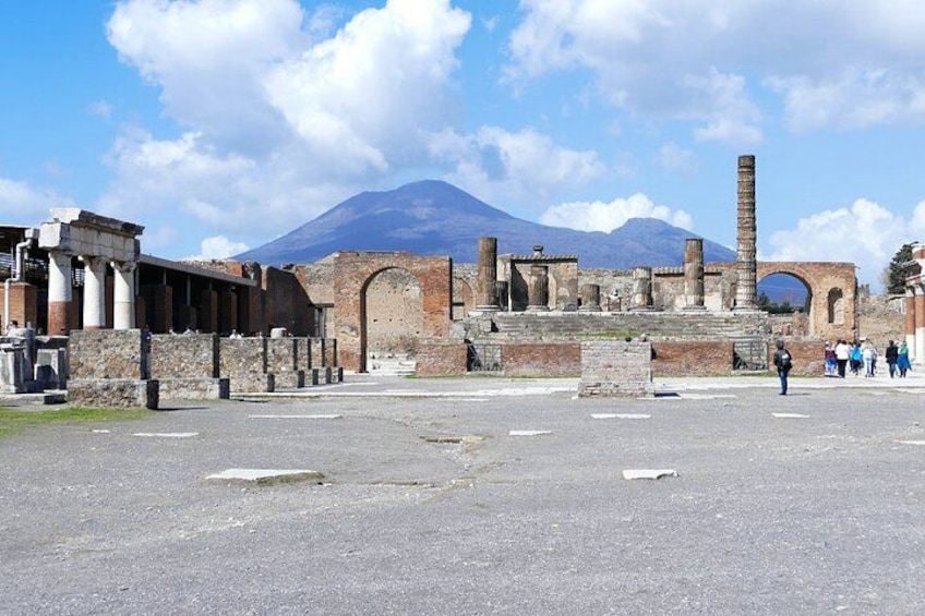 Ancient ruins with columns and arches, a volcano in the background, and tourists in the foreground. 1