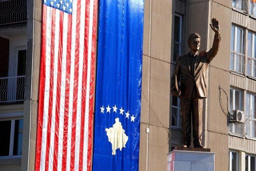 A statue of a man in a suit on a pedestal, with an American flag and a blue flag with stars on a building. 5