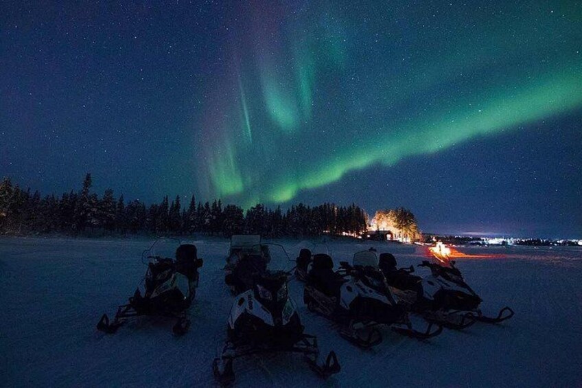 A snowy landscape with people on snowmobiles, a campfire, and the Northern Lights. 3