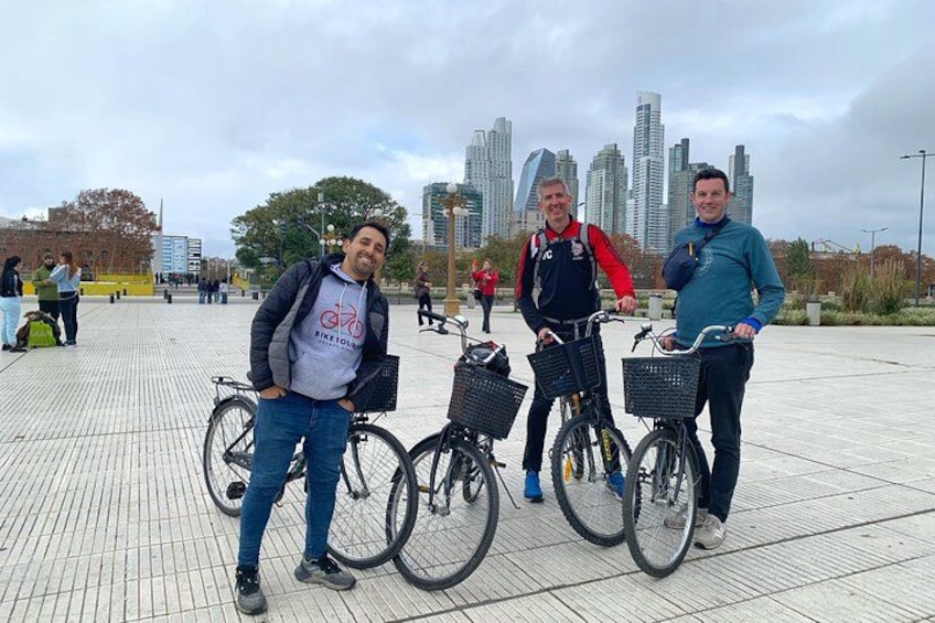 Three people standing next to bicycles on a paved area with a city skyline in the background. 3