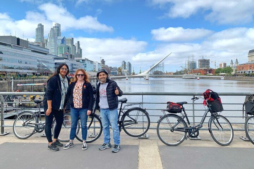 Three people standing by a river with bicycles, a city skyline, and a bridge in the background. 2