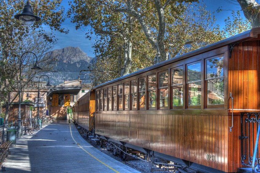 Un tren antiguo sobre rieles con un andén, árboles y montañas al fondo. 1
