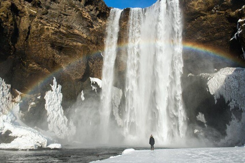 A waterfall with a rainbow, a person standing on ice, and snow-covered cliffs. 2