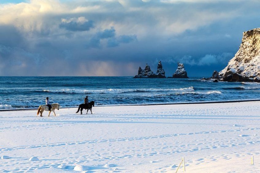 A snowy beach with horses and riders, ocean waves, and rocky formations. 3
