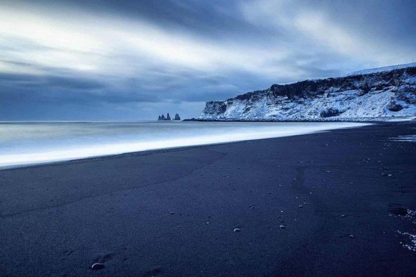 A beach with a dark sand shore, a calm sea, and a snow-covered cliff in the background. 5