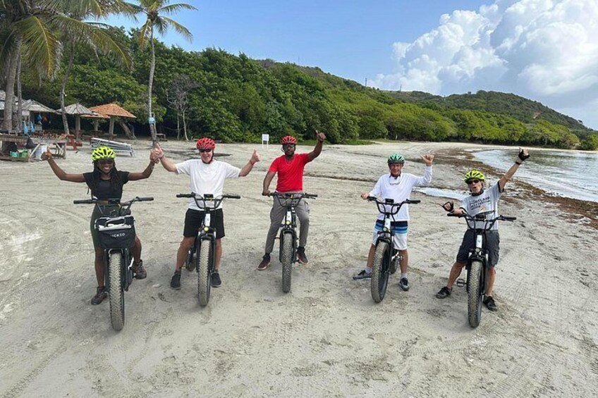 Five people on bicycles on a sandy beach with palm trees and a forested hill in the background. 1