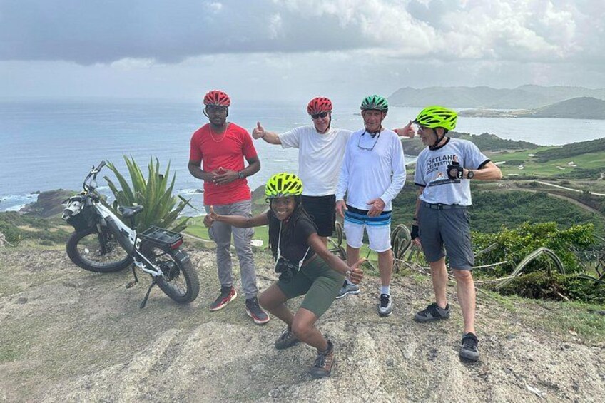 A group of cyclists on a coastal road with a view of the ocean and hills in the background. 3