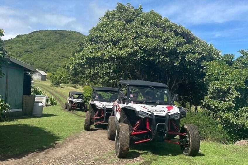 Two off-road vehicles parked on a dirt path in a rural area with greenery and a building in the background. 2