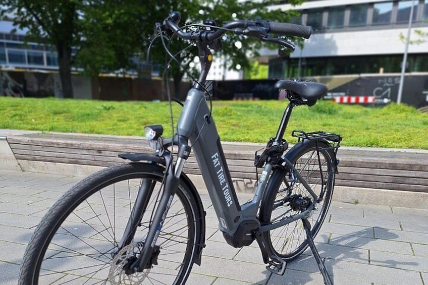 Un vélo électrique stationnaire, avec un cadre noir et un panier à l’arrière, est garé sur une surface pavée, avec de l’herbe et un bâtiment en arrière-plan. 2