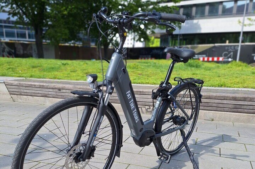 Een grijze elektrische fiets staat op een verhard oppervlak, met een grasveldje en een gebouw op de achtergrond. 2