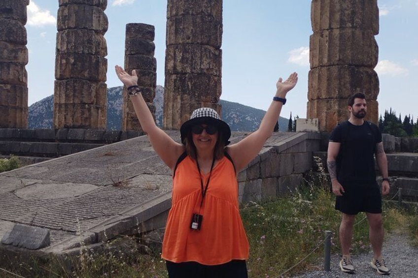 A person in an orange top and hat, standing in front of ancient stone columns with arms raised. 4