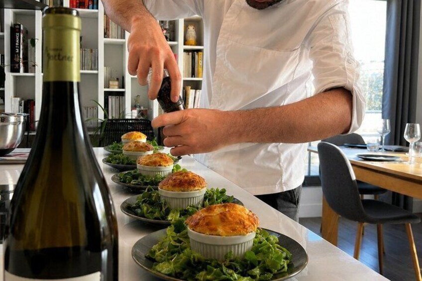 Un chef préparant des plats dans une cuisine, avec une bouteille de vin et des livres en arrière-plan. 1