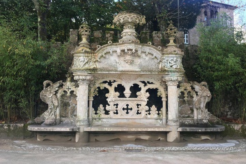 An ornate stone fountain with intricate carvings and a cross at its center, surrounded by lush greenery. 3