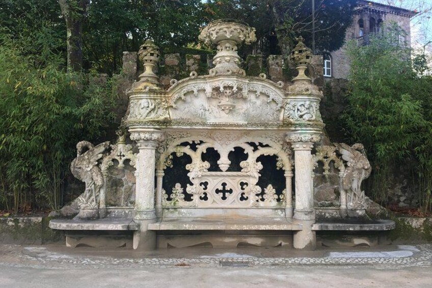 An ornate stone bench with intricate carvings, set against a backdrop of lush greenery and a historic building. 2