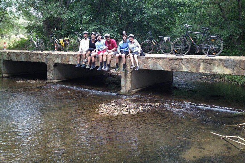A group of people sitting on a stone bridge over a stream, surrounded by trees. 4
