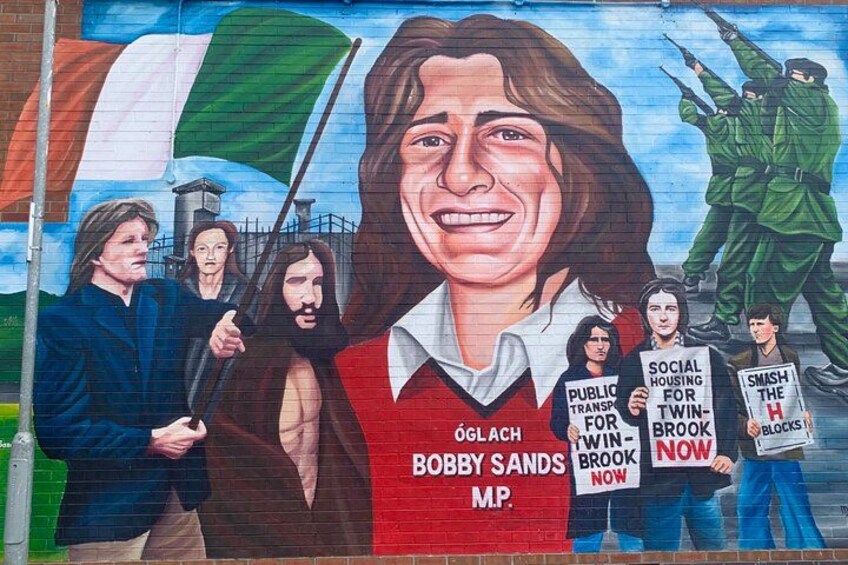Une fresque murale représentant une personne portant un gilet rouge sur lequel est inscrit « Bobby Sands, député », ainsi que plusieurs pancartes de protestation. 2