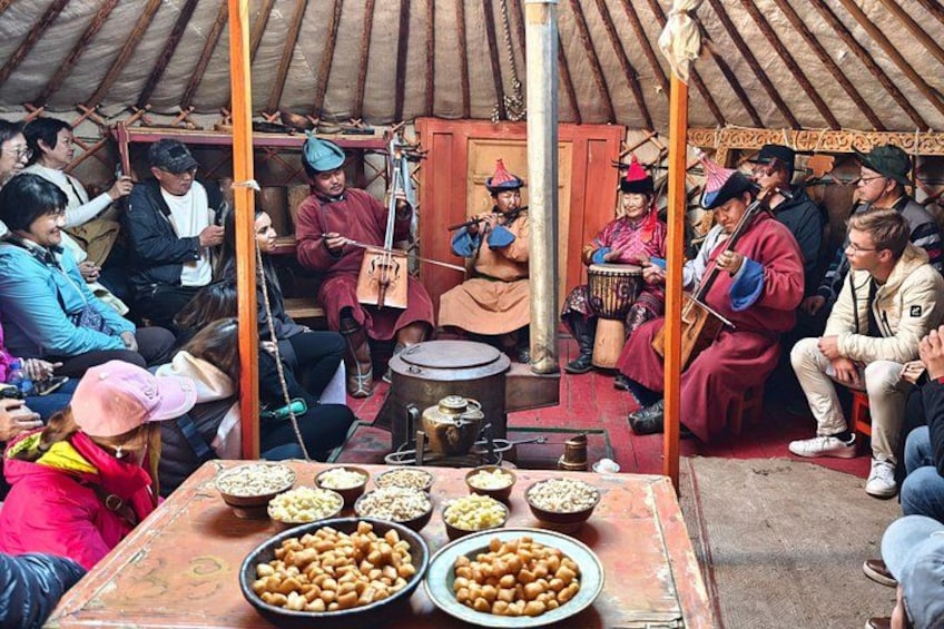 Een groep mensen zit rond een tafel met verschillende soorten eten en muziekinstrumenten, wat wijst op een gezellige bijeenkomst of evenement. 2