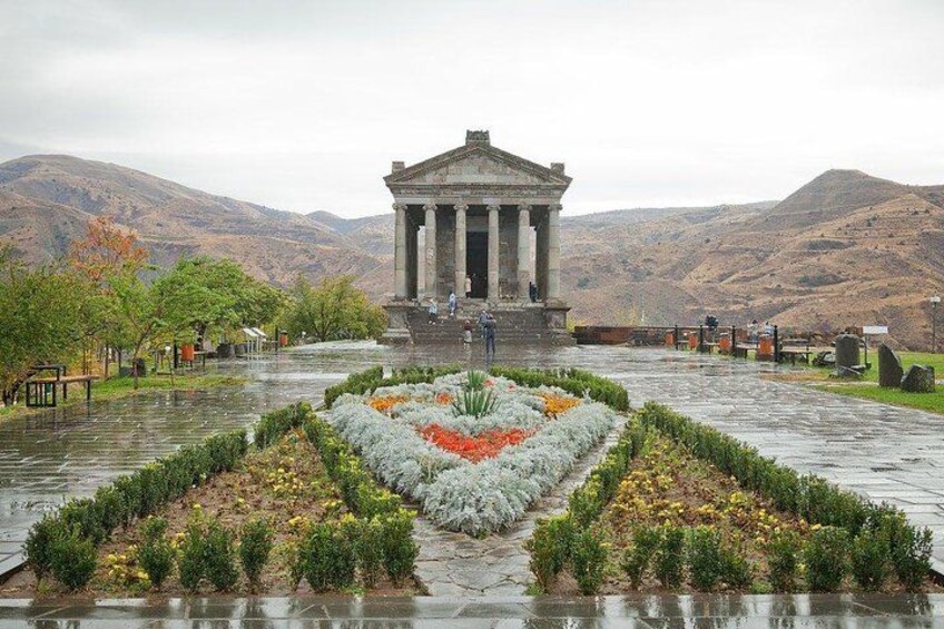 Ein klassizistisches Gebäude mit Säulen, umgeben von einem angelegten Garten mit einem zentralen Blumenbeet, vor dem Hintergrund von Bergen. 3
