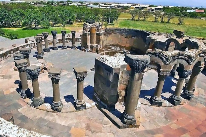 Columnas de piedra antiguas dispuestas en un círculo, con un bloque de piedra grande en el centro, frente a un fondo de vegetación y edificios. 2