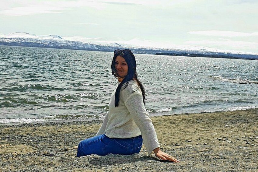 A person kneeling on a rocky shore with a body of water and snow-capped mountains in the background. 5