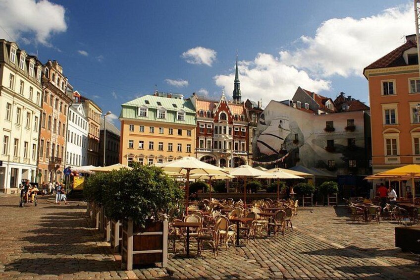 Une place pavée avec des tables en plein air, des bâtiments historiques et une grande fresque murale. 4