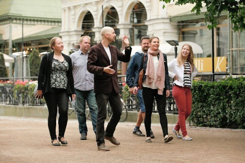 En gruppe mennesker går på en sti i et byområde med bygninger og en fontæne i baggrunden. 1