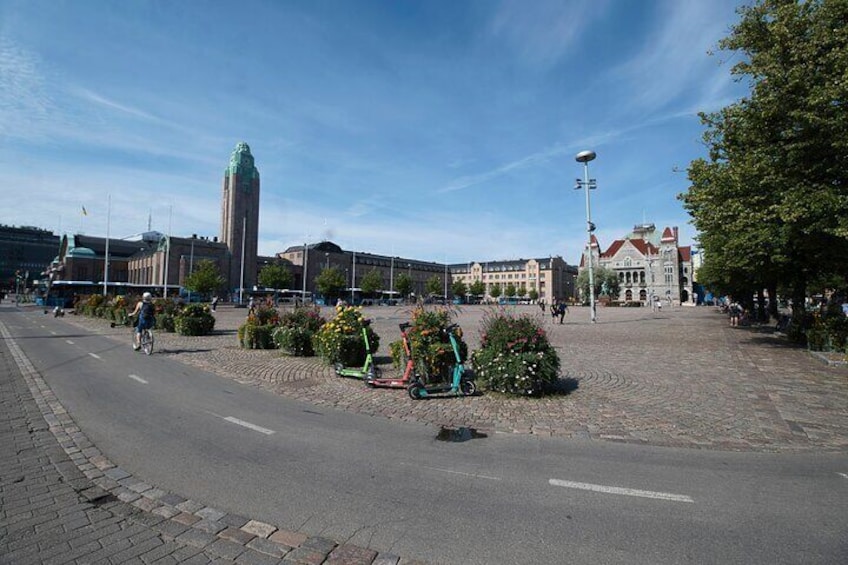 En anlagt plads med blomsterbed i midten, en høj bygning med grønt tag og en historisk bygning med røde og hvide vægge. 3