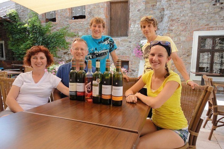 Een groep mensen zit aan een houten tafel met wijnflessen, in een rustieke buitenomgeving. 2