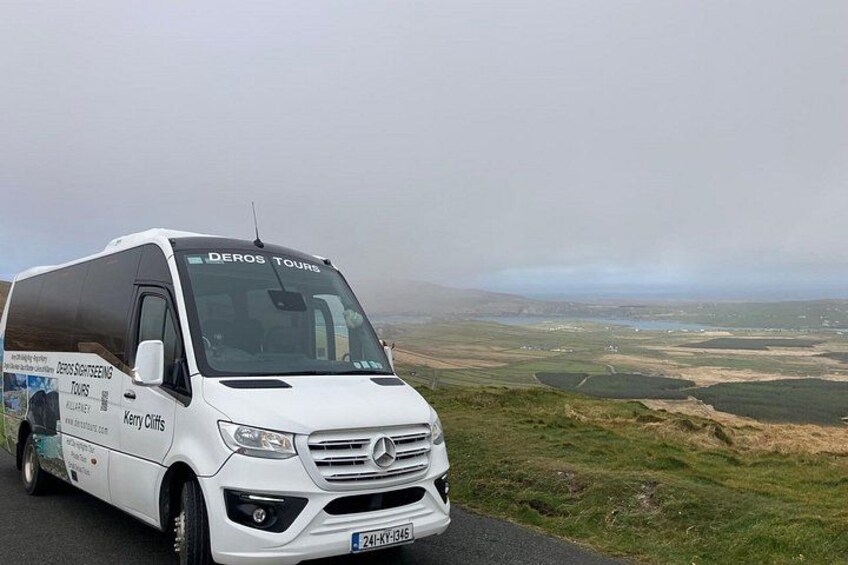 Una camioneta blanca Mercedes con la marca 'DEROS TOURS' y 'Kerry Cliff' estacionada en una carretera con una vista panorámica de colinas ondulantes y campos. 1