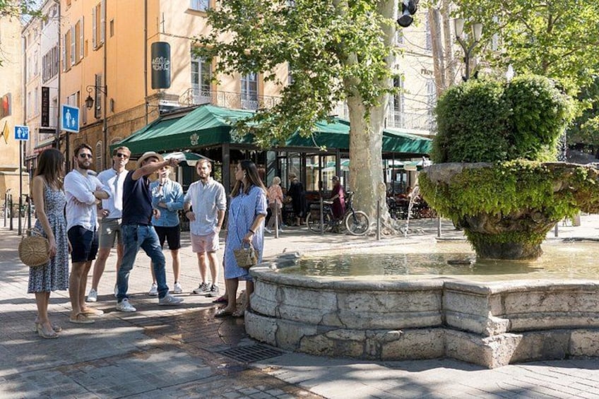 Un gruppo di persone in piedi attorno a una fontana in una piazza pubblica, con edifici e alberi sullo sfondo. 4
