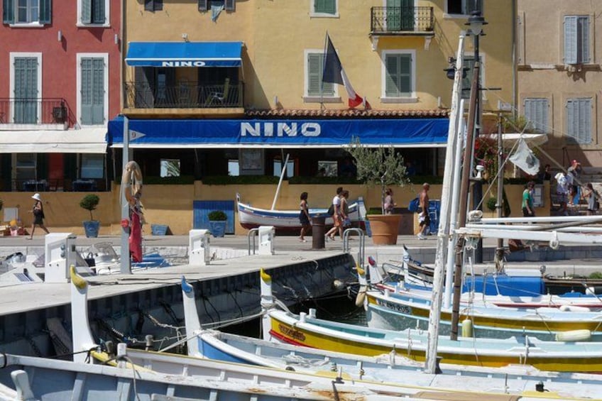 Una marina con diverse imbarcazioni ormeggiate, un ristorante NINO sullo sfondo e persone che passeggiano lungo il molo. 5