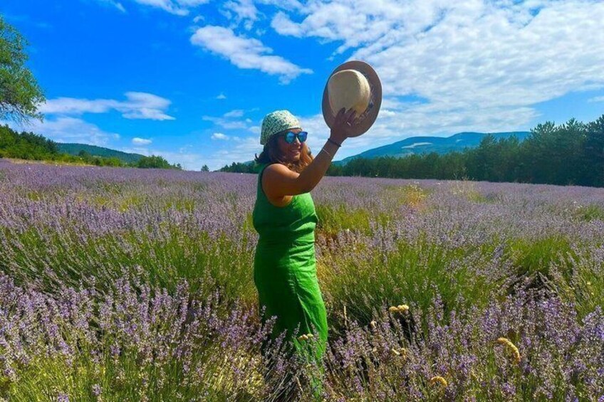 A person in a green dress holding a hat in a lavender field with mountains in the background. 3
