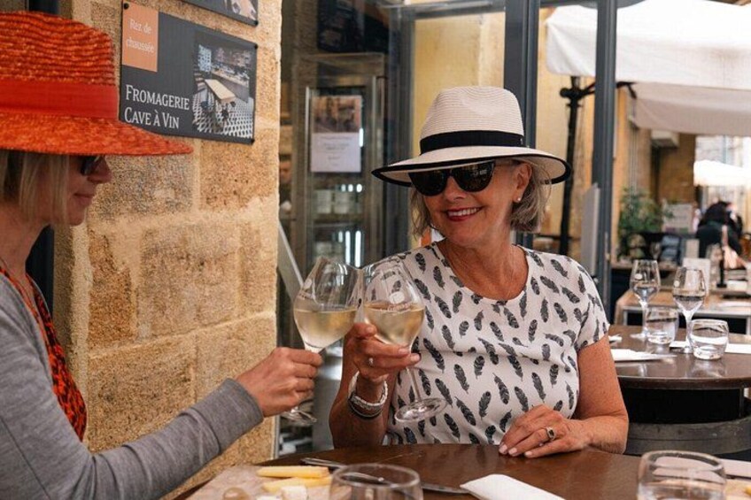 Two people toasting with wine glasses at a table in an outdoor setting with stone walls and a sign indicating a wine cellar. 4