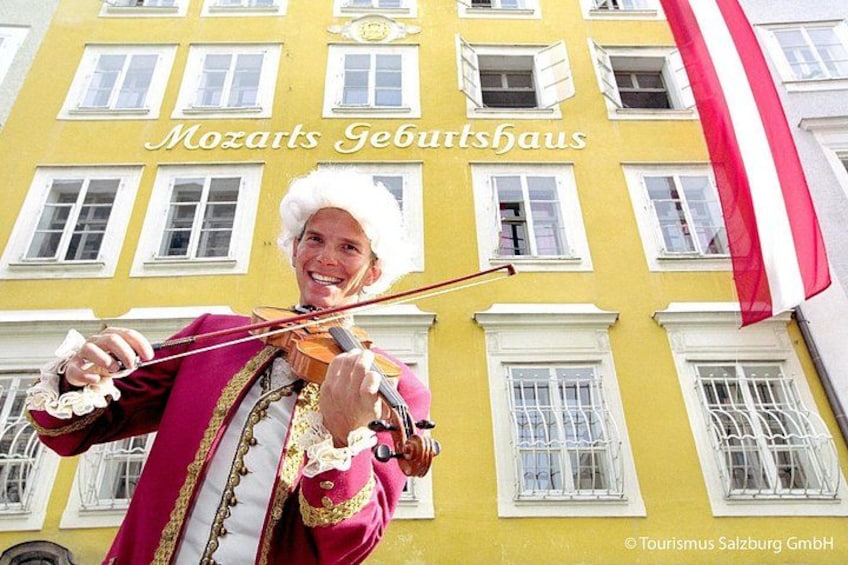 Eine Person spielt Violine vor einem gelben Gebäude mit dem Text 'Mozarts Geburtshaus'. 1