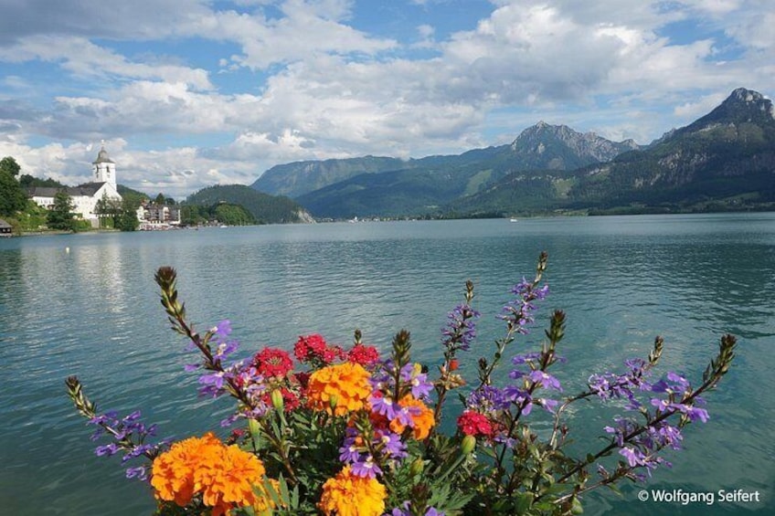 A lake with mountains in the background, a church on the left, and colorful flowers in the foreground. 4
