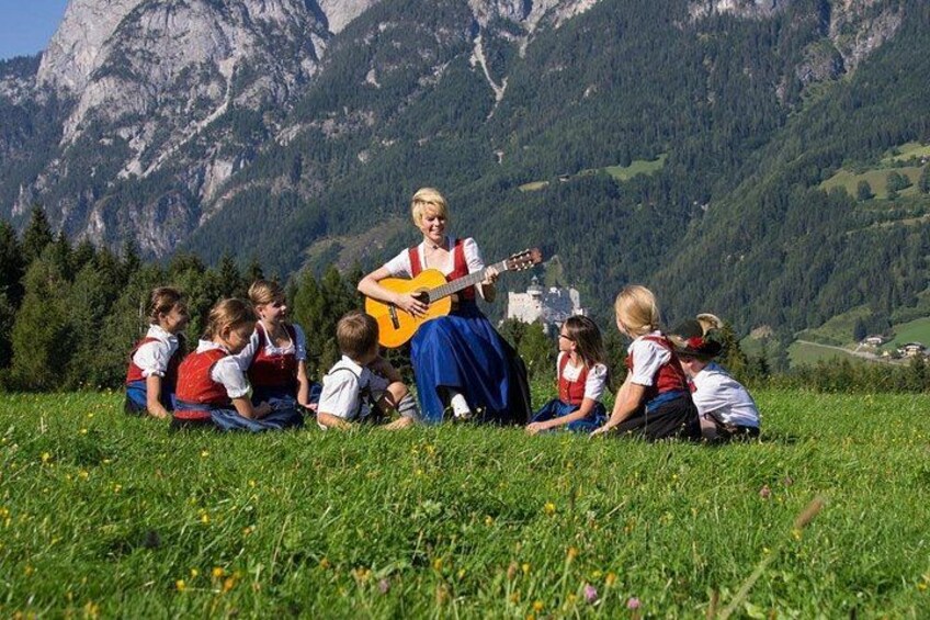 A woman playing guitar to a group of children outdoors with mountains in the background. 1