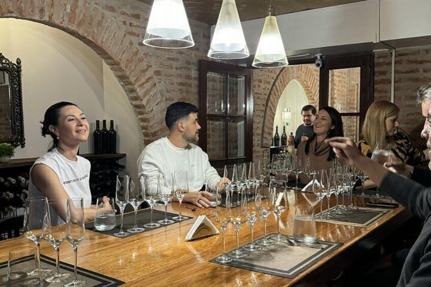 A group of people seated at a long wooden table with wine glasses and bottles, in a room with brick walls and hanging lights. 3