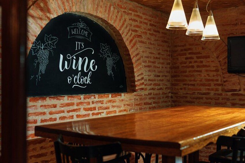 A brick wall with a chalkboard sign that reads 'Welcome, it's wine o'clock' and a wooden table with chairs. 4