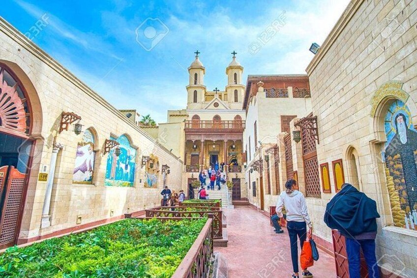 A church with a bell tower, a courtyard with plants, and people walking. 2