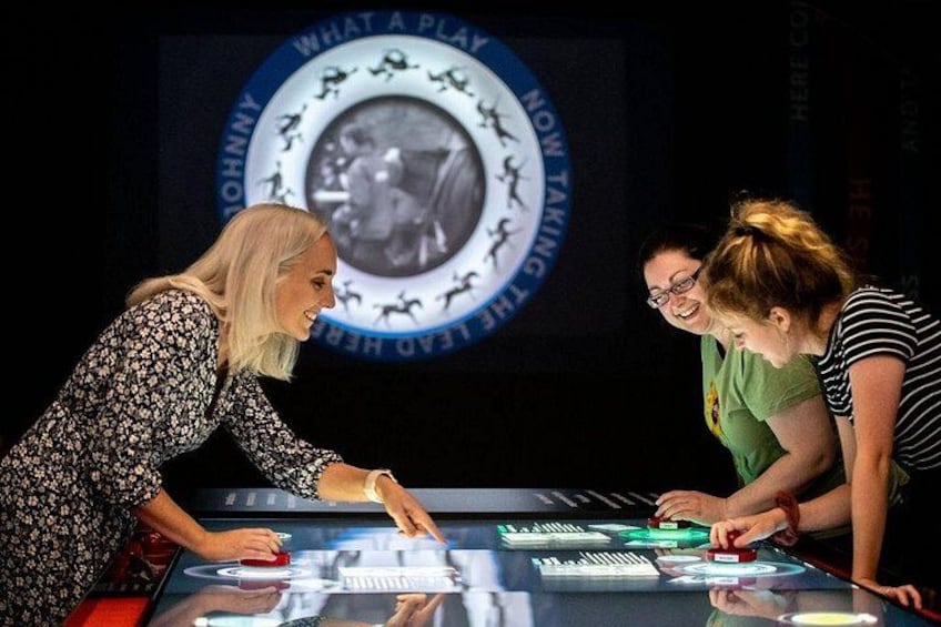 Three people interacting with a digital interface, possibly a game or interactive exhibit, with a circular emblem in the background. 2