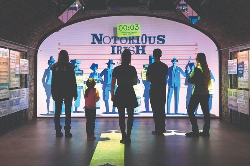 Un gruppo di persone all'interno di una struttura a forma di tunnel con un grande schermo digitale che mostra la scritta 'NOTORIOUS IRISH' e un orologio segna 00:03. 2