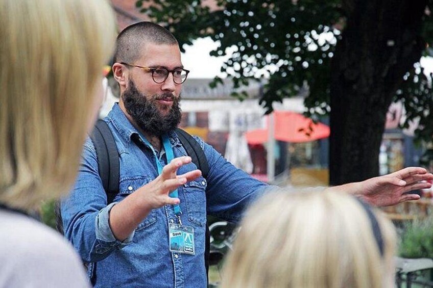 A person gesturing with hands, wearing a denim shirt and a lanyard, standing outdoors with trees and a building in the background. 2