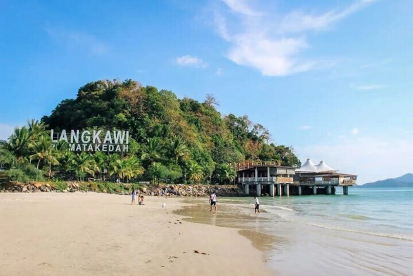 A beach with a large tree-covered hill, a building with a white roof, and calm water. 5